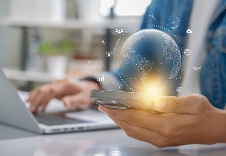 Person holding a smartphone with a glowing digital globe above it, sitting at a desk with a laptop in the background