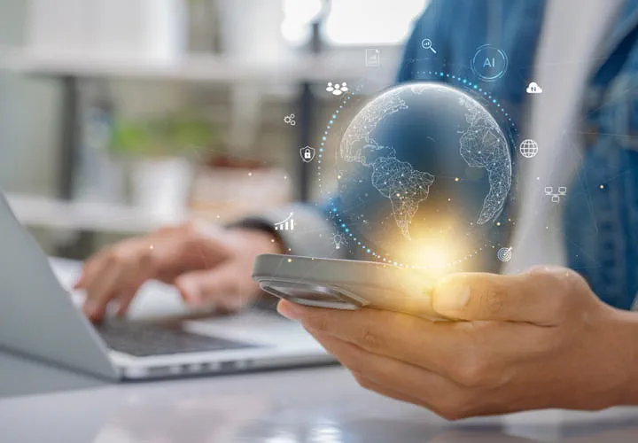 Person holding a smartphone with a glowing digital globe above it, sitting at a desk with a laptop in the background