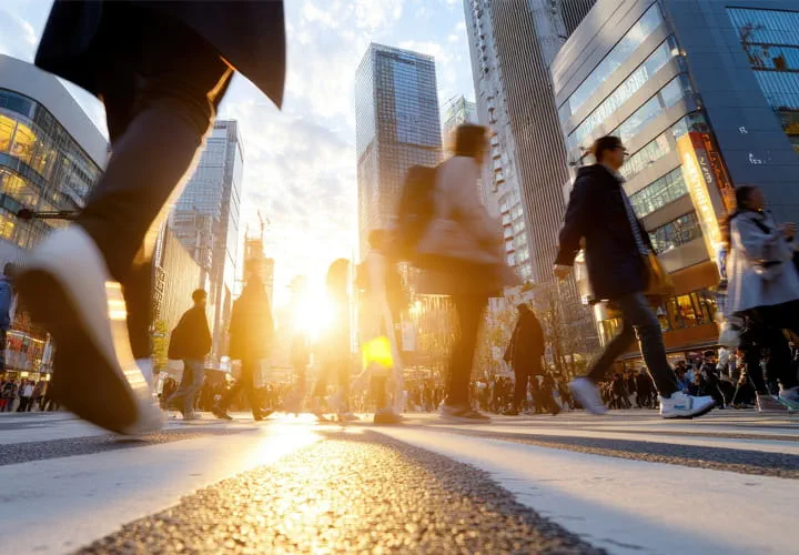 People crossing a city street with tall buildings and sunlight in the background